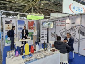 Image : Vue d'un stand de l'entreprise "smf" lors d'un salon professionnel, axé sur les solutions d'embouteillage, notamment pour les **bouteilles en plastique**. Le stand présente des panneaux d'affichage avec des images de diverses **bouteilles en plastique** remplies d'eau, de lait, d'huile comestible et de boissons gazeuses, ainsi que du texte sur les lignes d'embouteillage complètes pouvant atteindre jusqu'à 34 000 bph (bouteilles par heure). Au premier plan à gauche, un visiteur discute avec un représentant de l'entreprise. Au centre, un écran de télévision diffuse du contenu, et un autre représentant de l'entreprise est debout à côté d'une femme. Sur une table basse devant, plusieurs échantillons de bouteilles colorées sont exposés, ainsi que des brochures. Au premier plan à droite, deux hommes sont assis à une table, l'un en costume discutant avec l'autre, qui lui tourne le dos. Le stand est équipé de chaises et de tables blanches, et le sol est recouvert d'un tapis bleu. Au fond, une enseigne avec le logo "ACSR" est visible, et un panneau vert avec "Schneider". L'ensemble du stand met en avant l'expertise de smf en tant qu'**entreprise de bouteilles d'eau** et autres liquides.